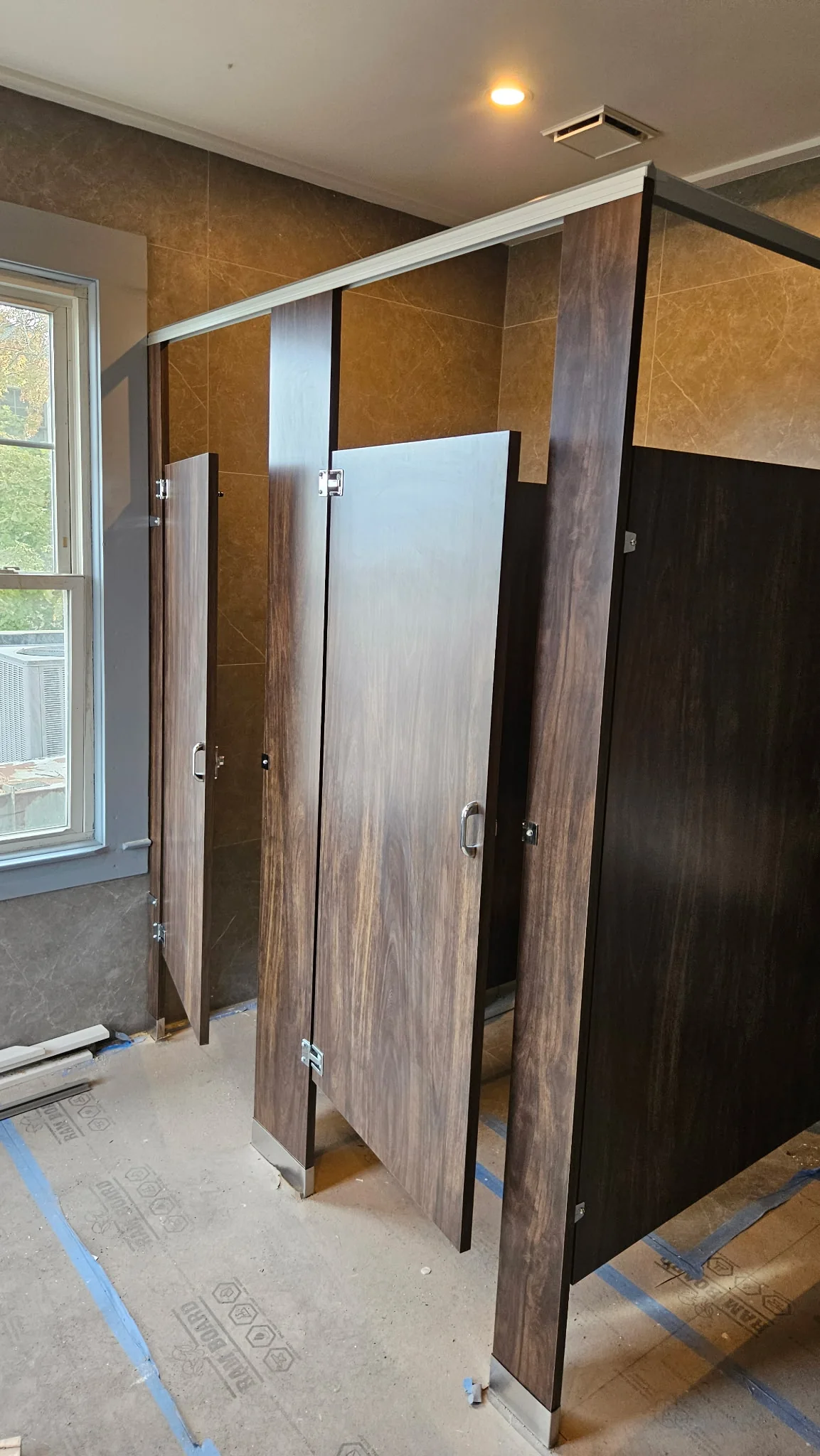 Wider installation-stage view of wood-look Plastic Laminate commercial restroom stall doors and partitions at La Maison in Greenwich, Connecticut